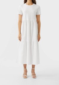 Femme portant une robe midi à manches courtes blanche unie avec un col rond et des talons blancs à lanières, se tenant contre un fond uni.