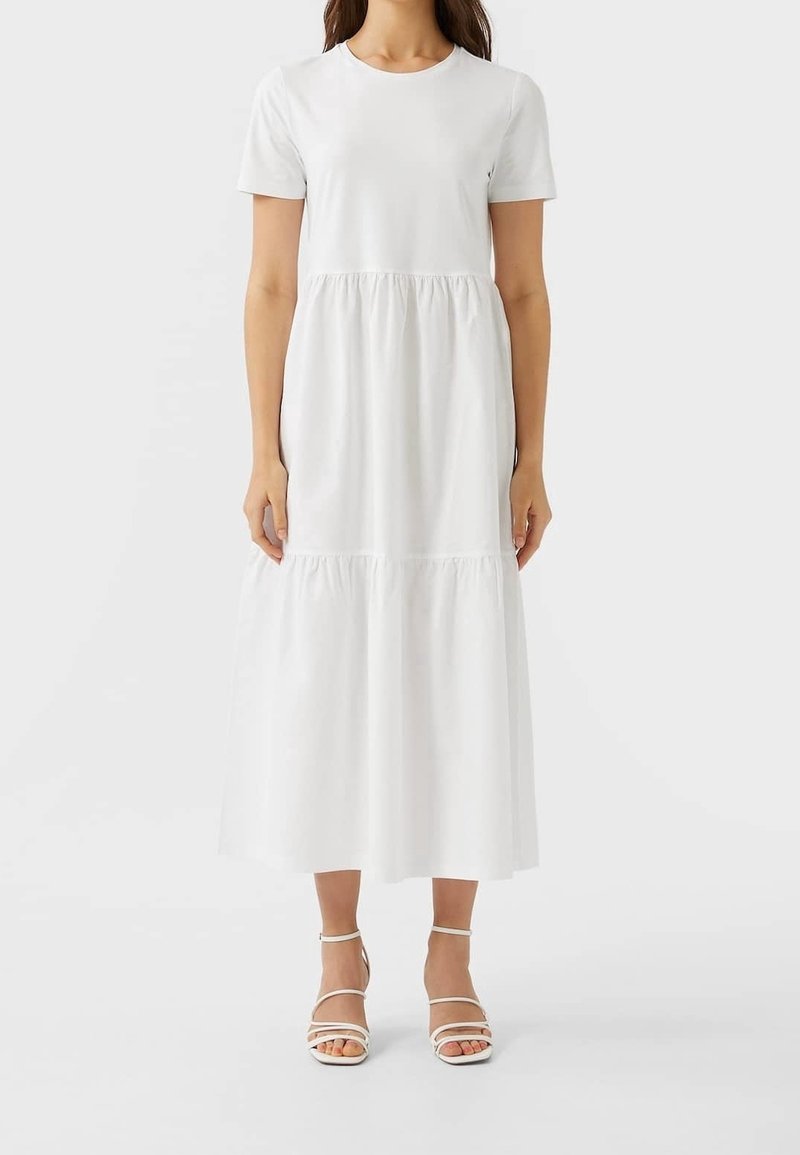 Femme portant une robe midi à manches courtes blanche unie avec un col rond et des talons blancs à lanières, se tenant contre un fond uni.