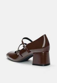Pantofi cu toc din piele patentă maro, cu vârful rotunjit, două curele ajustabile și un toc pătrat chunky. Textură netedă cu un finisaj lucios.
