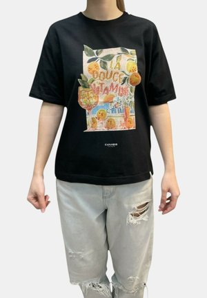 Persona che indossa una maglietta nera con frutti arancioni colorati e la scritta "La Douce Vitamine", abbinata a jeans grigi chiari strappati.