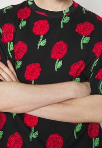 Svart t-shirt med ett mönster av röda rosor med gröna stjälkar, rund halsringning och korta ärmar. Mjuk bomullstextur.
