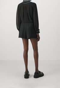 Blouse noire transparente à manches longues, short noir plissé et chaussures à plateforme noires, avec un col et des poignets décoratifs.