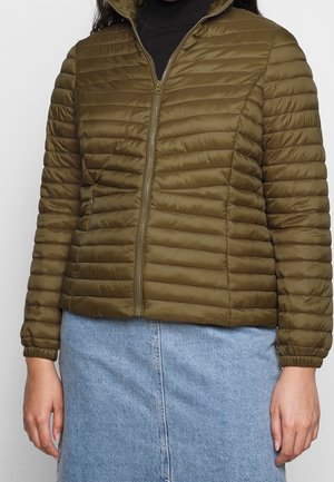 Femme portant une veste matelassée vert olive avec une fermeture éclair à l'avant et une jupe en denim bleu clair, debout devant un fond uni.