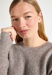 Žena s dlouhými blond vlasy opírá bradu o ruku a má na sobě světle hnědý svetr, na neutrálním pozadí.