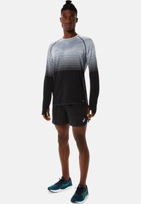 Langarm sporttop in gradiëntgrijs en zwart, gecombineerd met zwarte shorts, gedragen met blauw-groene hardloopschoenen. Bevat logo-accenten.