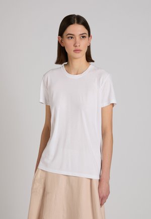 Mujer joven con cabello castaño lacio, viste una camiseta blanca de manga corta y una falda beige, mirando ligeramente hacia la derecha.