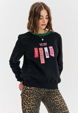 Czarna bluza z zielonym kołnierzykiem, na której umieszczone są kolorowe metki z napisami "MIŁOŚĆ", "RÓWNOWAGA", "RADOŚĆ", "RYZYKO" oraz "Weź, czego potrzebujesz."