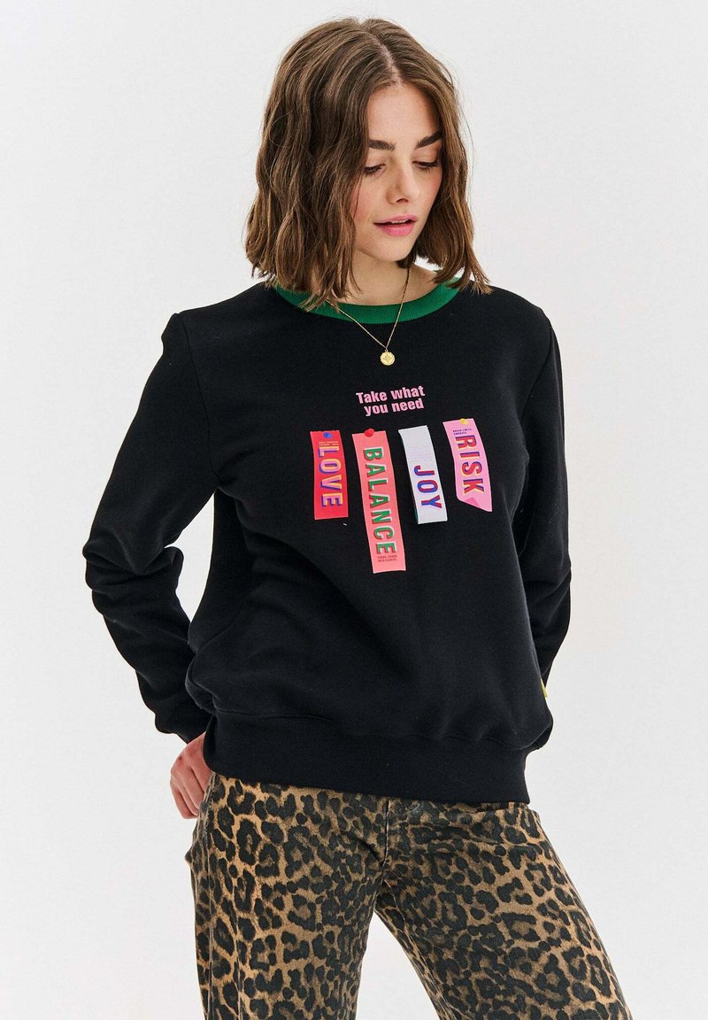 Černá mikina s zeleným límečkem obsahuje barevné textilní štítky s nápisy "LÁSKA", "ROVNOVÁHA", "RADOST", "RIZIKO", a "Vezmi, co potřebuješ."