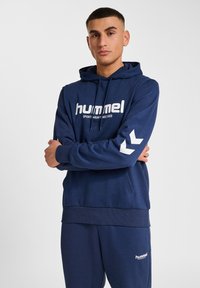 Granatowa bawełniana bluza z kapturem i sznurkiem, z białym logo "hummel" oraz akcentami w kształcie chevronu na rękawach. Zestawiona z pasującymi spodniami.