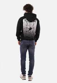 Felpa nera con un design grafico di costole e un effetto frantumato sul retro. Abbinata a jeans blu e scarpe da ginnastica chiare.