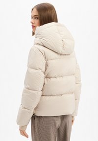 Woman with straight hair wears a light beige puffer jacket with large hood, paired with light brown checkered pants, facing left with head turned.