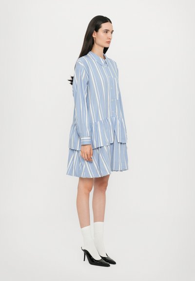 Blau-weiß gestreiftes Hemdkleid mit langen Ärmeln, einer durchgehenden Knopfleiste und einem gestuften Design. Kombiniert mit weißen Socken und schwarzen High Heels.