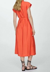 Koronkowa sukienka midi w kolorze koralowym z krótkimi rękawami, paskiem w talii i teksturowanym materiałem. Zestawiona z czarnymi sandałami na cienkich paskach. Widok z tyłu.
