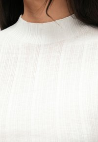 Λευκό ριμπ υψηλόλαιμο τοπ με υφασμένο μοτίβο και εφαρμοστή σιλουέτα. Το ύφασμα φαίνεται μαλακό και ελαφρύ με διακριτική λάμψη.