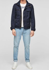 Mörkblå vadderad jacka med ståkrage, frontdragkedja och flera fickor, kombinerad med ljusblå jeans och vita sneakers.