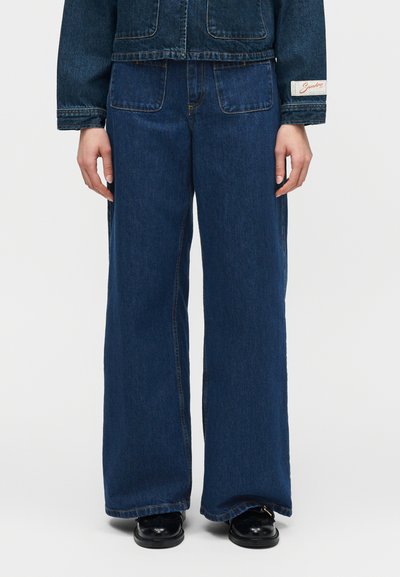 Άτομο που φοράει τζιν παντελόνι με φαρδιά μπατζάκια σε σκούρο μπλε και τζιν μπουφάν στο ίδιο χρώμα με εμφανείς μπροστινές τσέπες και μαύρα παπούτσια.