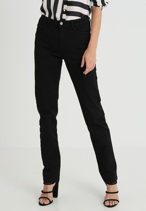 Persoon die zwarte slim-fit jeans, zwarte sandalen met hoge hakken en bandjes, en een zwart-wit gestreept shirt draagt, staand tegen een effen achtergrond.