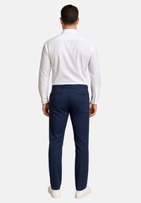 Weißes Langarmhemd und marineblaue Hose im Slim-Fit, mit glatter Textur, seitlichen Taschen und zwei Knöpfen auf der Rückseite. Weiße Sneaker runden den Look ab.