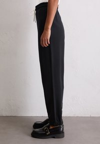 Zwarte sweatpants met een relaxte pasvorm, een trekkoord in de taille en elastische boorden. Soepele textuur met minimale details. Gepaard met zwarte loafers.