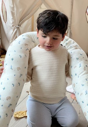 Jeune enfant en pull beige tenant un grand coussin blanc à motifs floraux, assis sur un sol en bois près d'une tente de jeu en tissu.