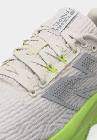 Κοντινό πλάνο λευκού και ανοιχτό πράσινου παπουτσιού τρεξίματος New Balance με υφασμάτινη υφή, ετικέτα στη γλώσσα με το λογότυπο και πλεκτά κορδόνια περασμένα από τρύπες.