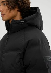 Chaqueta de plumas negra con cierre de cremallera, capucha ajustable y tejido texturizado. Presenta un parche con el logo de ellesse en la manga.