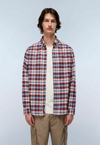 Giovane in piedi, che indossa una camicia a quadri rossa, bianca e blue con bottoni sopra una maglietta bianca e pantaloni cargo beige, su uno sfondo neutro.