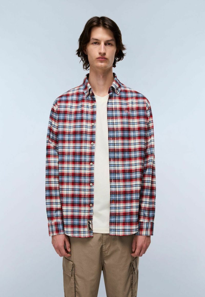 Giovane in piedi, che indossa una camicia a quadri rossa, bianca e blue con bottoni sopra una maglietta bianca e pantaloni cargo beige, su uno sfondo neutro.