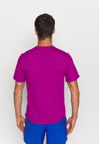 Hombre con pelo corto y oscuro que lleva una camisa de manga corta de color púrpura brillante y pantalones cortos azules, de espaldas contra un fondo blanco liso.