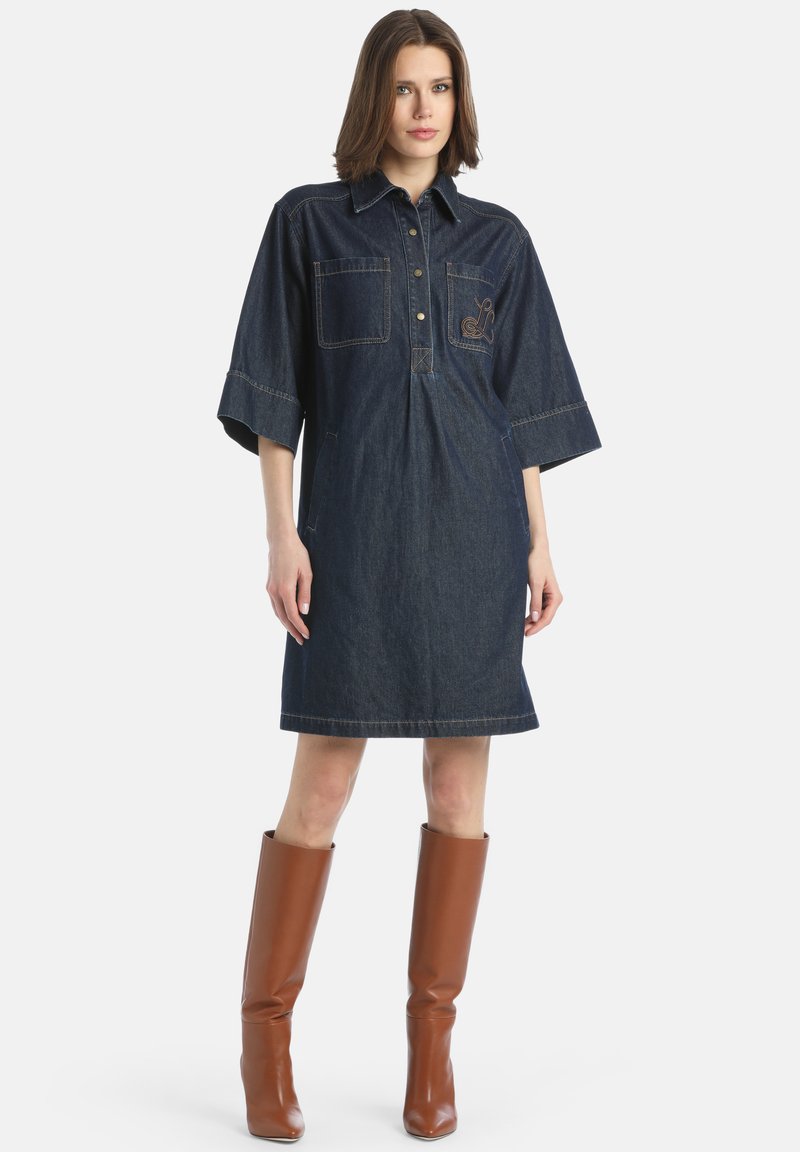 Denim-paitamekko, jossa on lyhyet hihat, nappikiristys etuosassa, kaksi rintataskua ja vaaleanruskea brodeeraus yksityiskohtana; yhdistettynä pitkään ruskeaan saappaaseen.