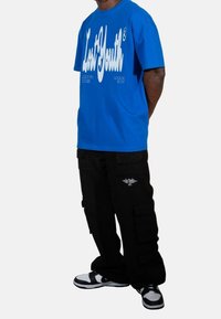 Sininen puuvillapaita, jossa on valkoinen graafinen teksti, yhdistettynä mustiin cargo-housuihin, joissa on sivutaskut ja logo, sekä musta-valkoisiin lenkkareihin.