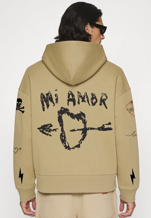 Person trägt beige Kapuzenjacke mit dem Text "Mi Amor" und einem Herz, das von einem Pfeil durchbohrt ist, auf dem Rücken, Totenkopf- und Blitzsymbole auf den Ärmeln.