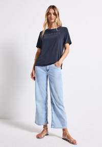 Námořnické tričko s krátkým rukávem a tištěným textem, spárované se světle modrými širokými kalhotami a stříbrnými sandály na špagetové pásky.