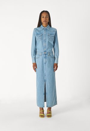 Abito camicia in denim azzurro chiaro con maniche lunghe, due tasche sul petto e un'apertura frontale. Indossato con sandali gialli con cinturini.