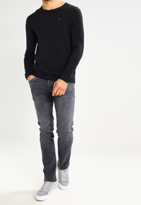 Haut noir à manches longues et col rond associé à un jean gris slim et des baskets grises. Logo brodé remarquable sur la poitrine. Texture lisse.