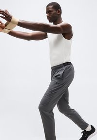 Άνδρας με λευκή αμάνικη μπλούζα και γκρι παντελόνι Under Armour τεντώνει τα χέρια προς τα εμπρός με λάστιχο αντίστασης, ισορροπώντας σε ένα πόδι.