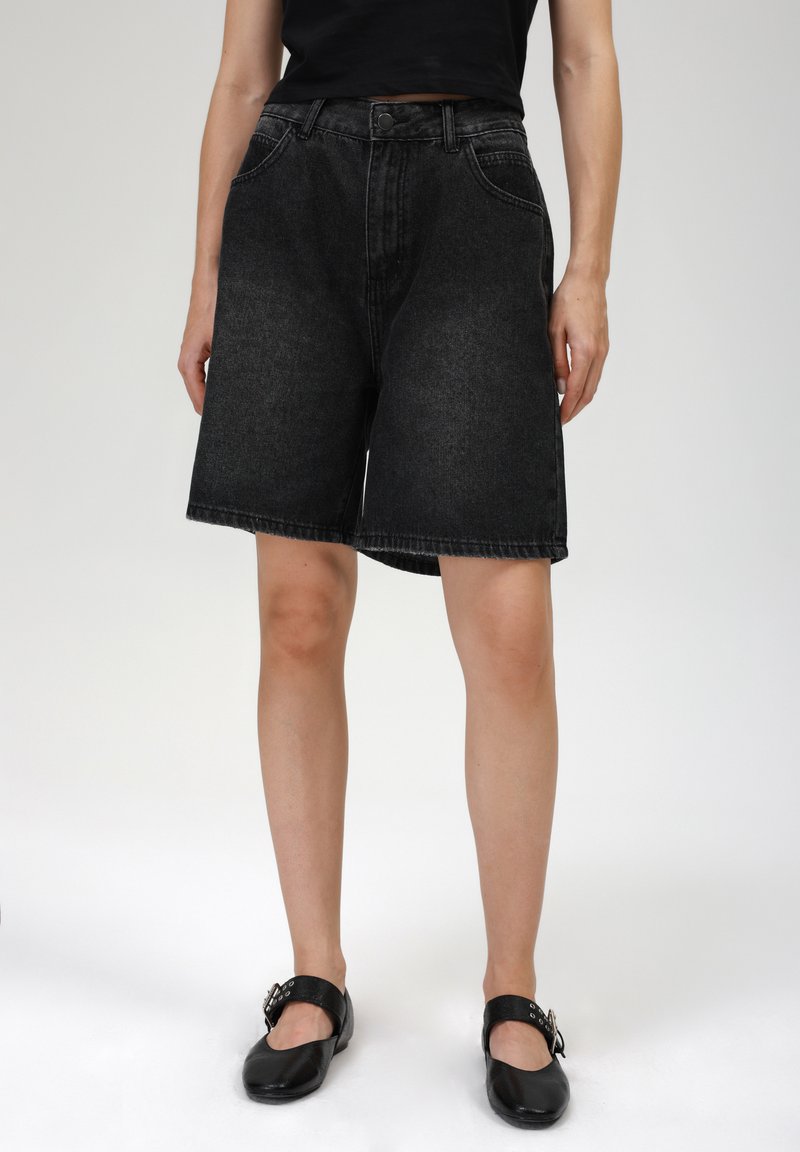 Shorts en denim noir avec une coupe ample, poches avant et taille haute. Longueur courte avec un ourlet brut. Associés à des chaussures plates noires.