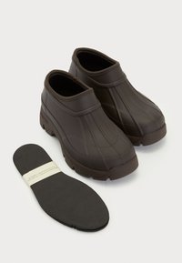Saboti din cauciuc maro cu un design texturat, talpă joasă și talpă groasă. Include o branț detașabil negru.