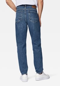 Jeans de mezclilla azul con corte recto, que cuentan con bolsillos traseros y una etiqueta de marca de cuero marrón en la cintura. Llevados con zapatillas blancas.