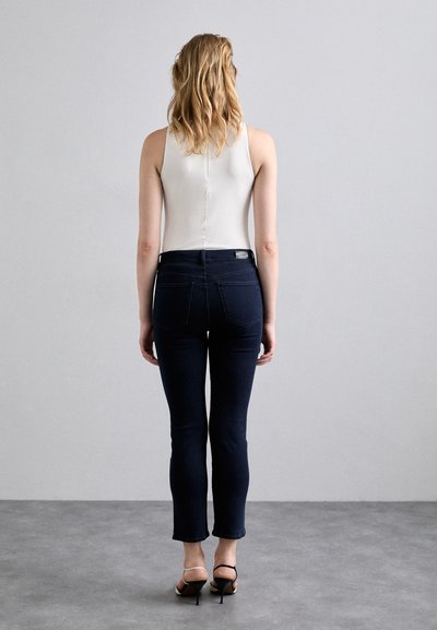 Jean skinny noir à taille mi-haute, avec des poches arrière et un ourlet légèrement court, accompagnés d'un haut sans manches blanc et de sandales à talons.