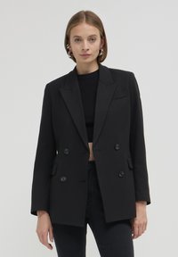 Blazer nero doppiopetto con rever affilati, due tasche frontali e bottoni neri rotondi. Realizzato in tessuto strutturato con una texture liscia.