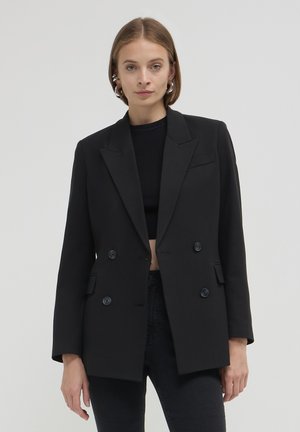 Blazer noir croisé avec des revers accentués, deux poches avant et des boutons ronds noirs. Fabriqué en tissu structuré avec une texture lisse.