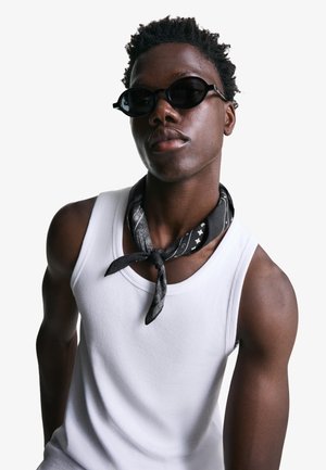 Giovane uomo con occhiali da sole neri, canotta bianca e foulard nero a motivo intorno al collo, su sfondo chiaro uniforme.