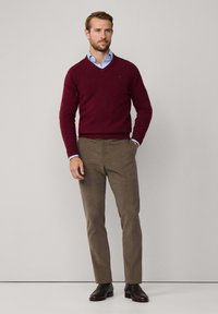 Pull à col en v bordeaux, chemise à col bleu clair en dessous, pantalon kaki, chaussures habillées marron, debout sur un fond neutre.