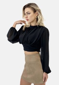 Schwarze, bauchfreie Bluse mit gerafften Details und langen, transparenten Ärmeln, kombiniert mit einem eng anliegenden, beigen Minirock aus Wildleder. Goldene Ohrringe runden den Look ab.