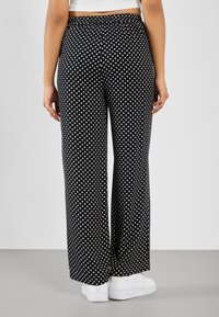 Pantalon large à pois noirs avec une ceinture élastique, en tissu léger, associé à des baskets blanches dans un cadre minimaliste.