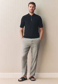 Črna polo majica s kratkimi rokavi in zadrgo, kombinirana s svetlo sivimi ohlapnimi hlačami in črnimi natikači, stoji ob gladki steni.