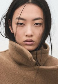 Cappotto marrone di lana con collo alto, texture morbida e design pulito e minimalista. Il modello ha capelli lunghi neri con ciocche sciolte.