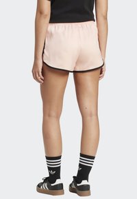 Světle růžové sportovní šortky s černým lemováním, vybavené elastickým pasem a rozštěpeným lemem pro lepší pohyb. Kombinovány s černými teniskami a ponožkami.