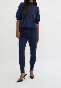 Námořnické modré, volné tričko s nabíranými rukávy, spárované s odpovídajícími tepláky. Hladká textura; doplněné černými špičatými podpatky.